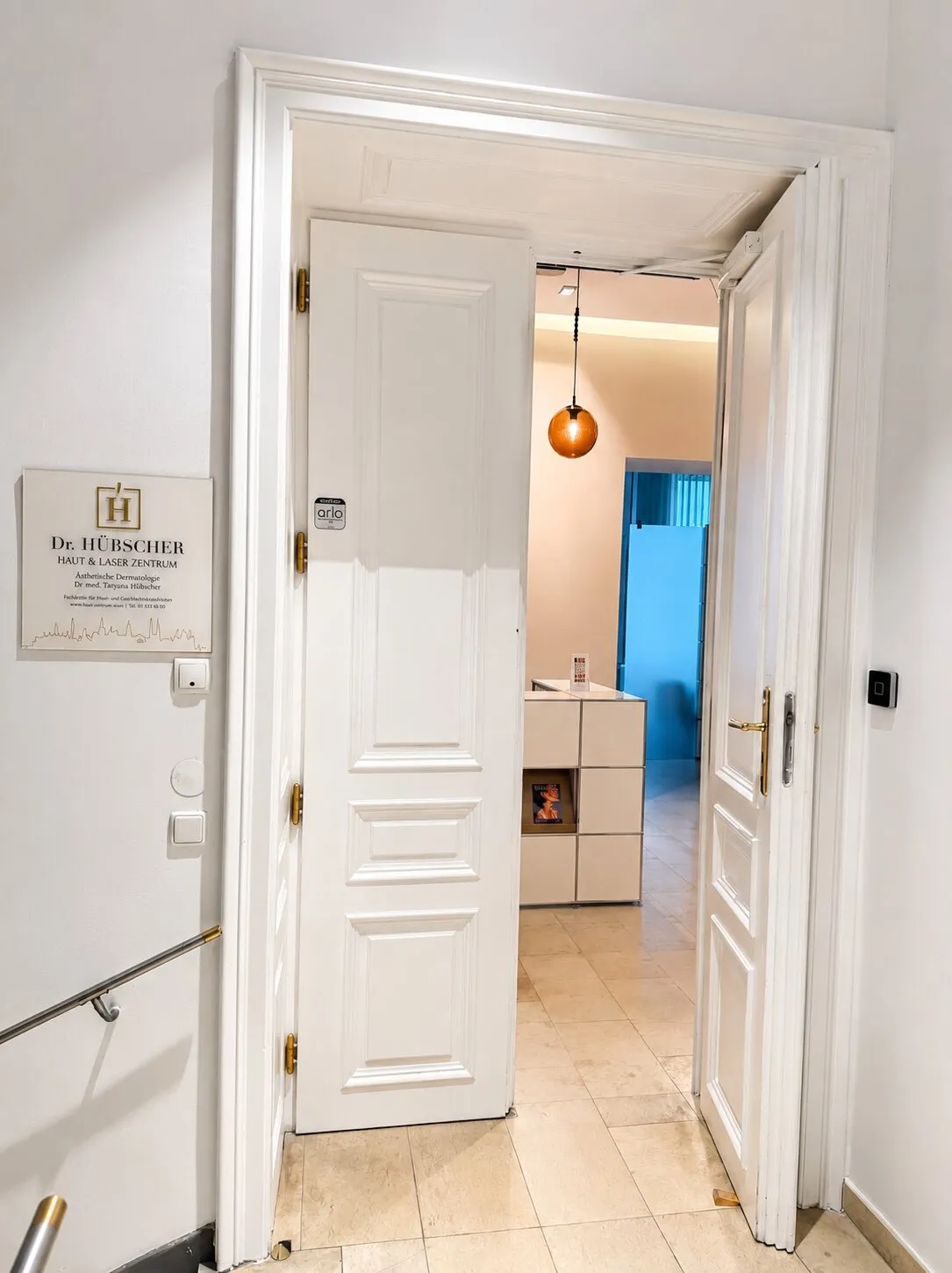 Practice entrance with an open white double door, the Dr. Hübscher practice sign and a view through to reception