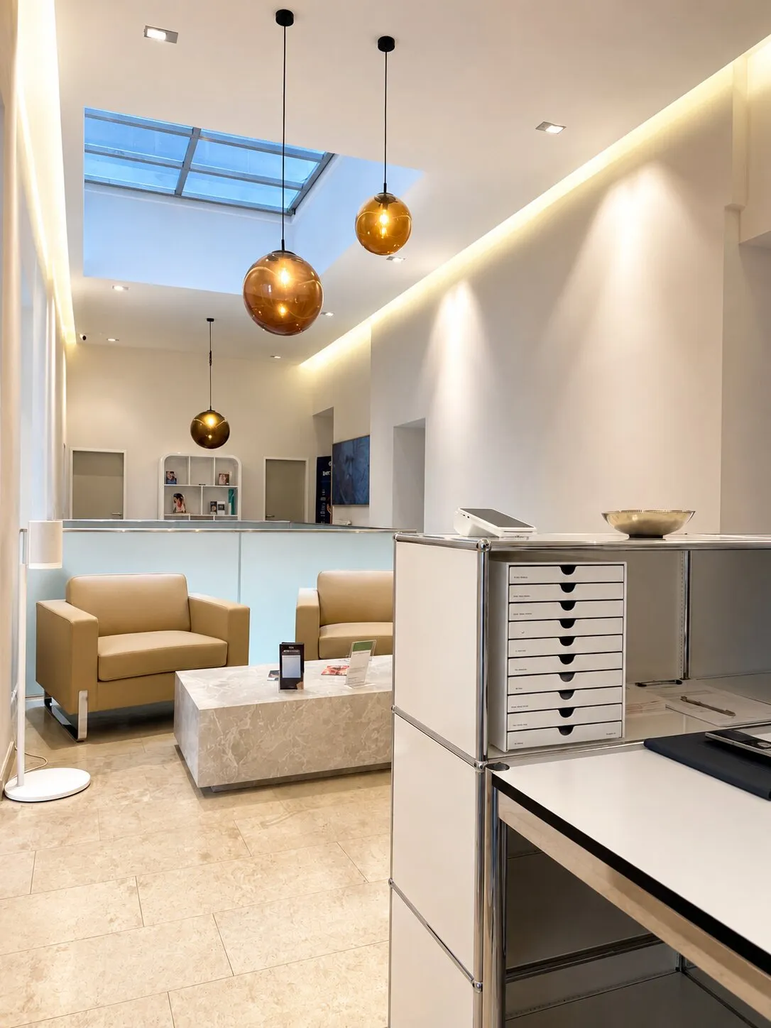 Waiting area of the practice with light leather armchairs, a marble table and amber pendant lamps beneath a skylight